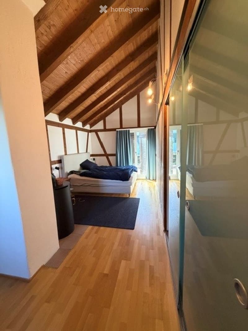 Cozy attic bedroom with wooden slanted ceilings, exposed beams, and hardwood flooring. Features a white bed with blue bedding, a nightstand, and a dark blue rug.