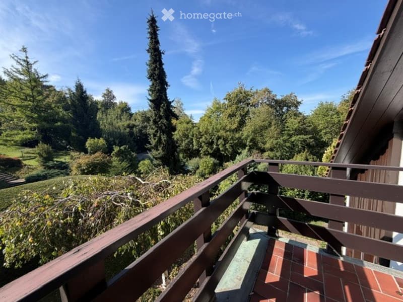 Balcony with wooden railing, overlooking a lush green garden and tall trees under a clear blue sky.
