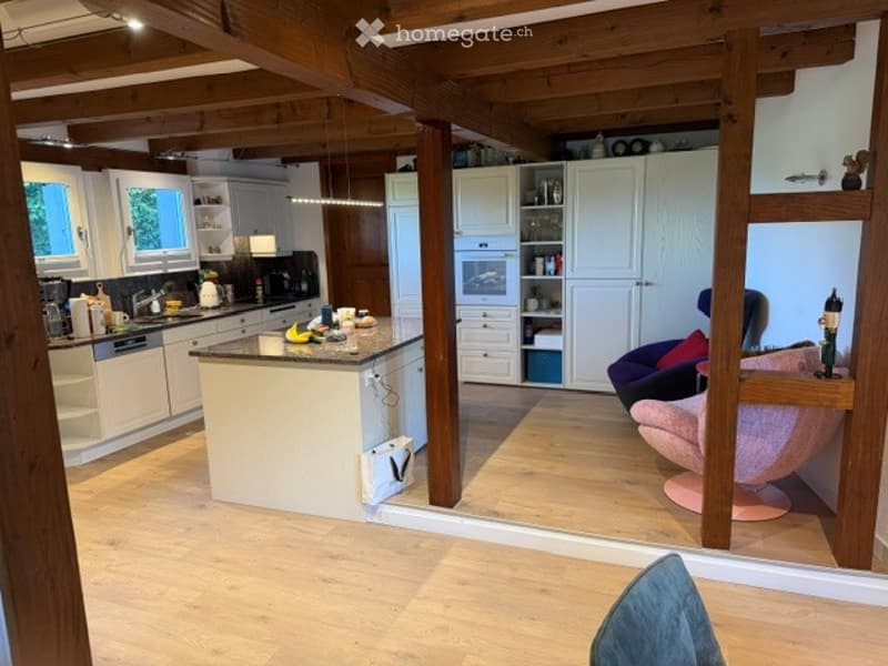 Open-plan kitchen and living area with white cabinets, a central island, and modern appliances. Wooden beams on the ceiling and a pink chair near the island.