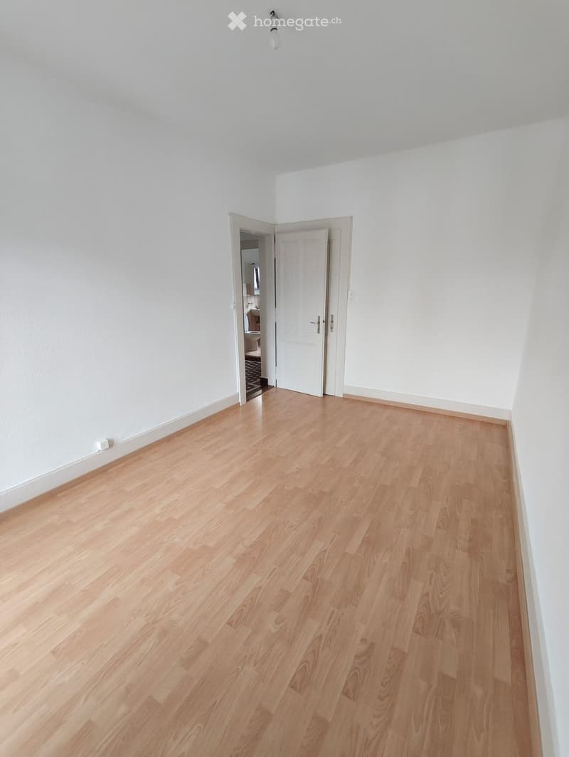 Empty room with light wooden flooring and white walls, featuring an open doorway leading to another room.
