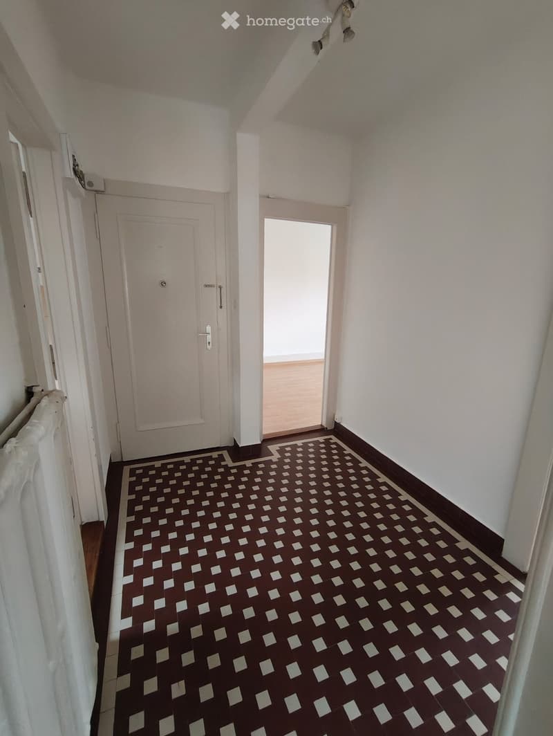 A narrow hallway with patterned tiles, white walls, and a closed door on the left and an open doorway on the right.
