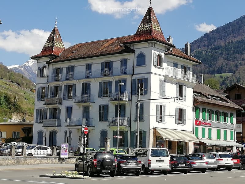 A large, white, multi-story building with blue shutters and balconies, featuring a distinctive red-tiled roof and ornate architectural details. The building is situated in a scenic area with mountains in the background and several parked cars in front.
