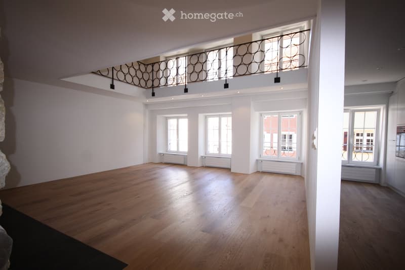 Large empty room with wooden floor, white walls, three windows with radiators, black railing on raised ground floor