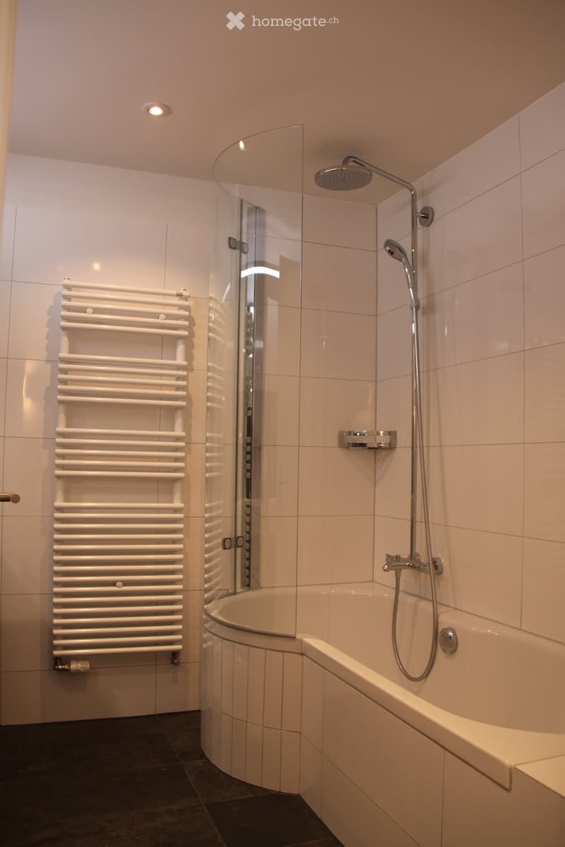 white tile walls, corner bathtub, glass shower enclosure, shower head, towel heater