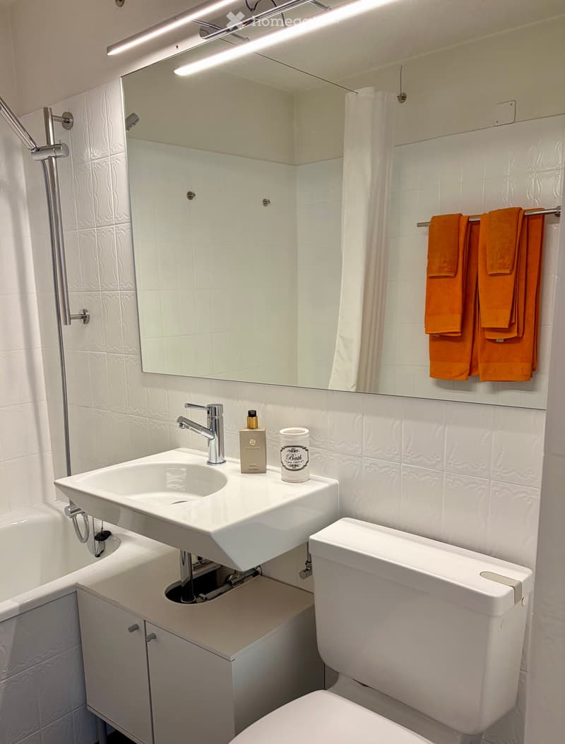 Modern bathroom with white tiles, a large mirror, a sink with a modern faucet, a toilet, a bathtub, and a towel rack with orange towels.