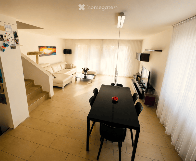 Bright living and dining area with a white sofa, coffee table, dining table with chairs, TV, and a cat tree. Large windows with white curtains allow natural light to fill the space.
