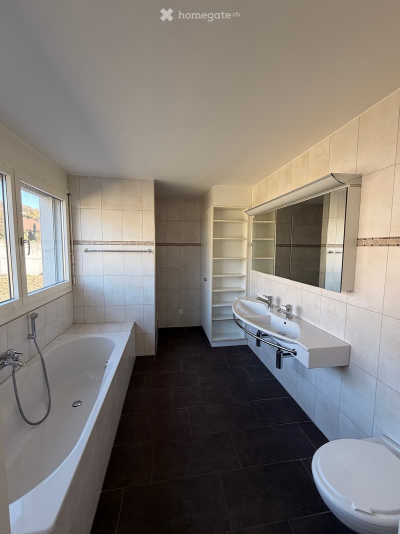 Modern bathroom with a bathtub, double sink, toilet, mirror, and built-in cabinet. Dark tiled floor and white tiled walls.