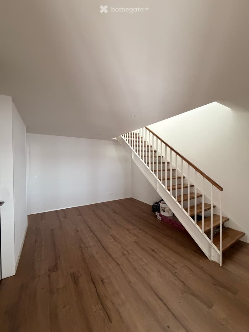 Empty hallway with wooden flooring, white walls, and a staircase leading upwards. The staircase features wooden steps and white railings.