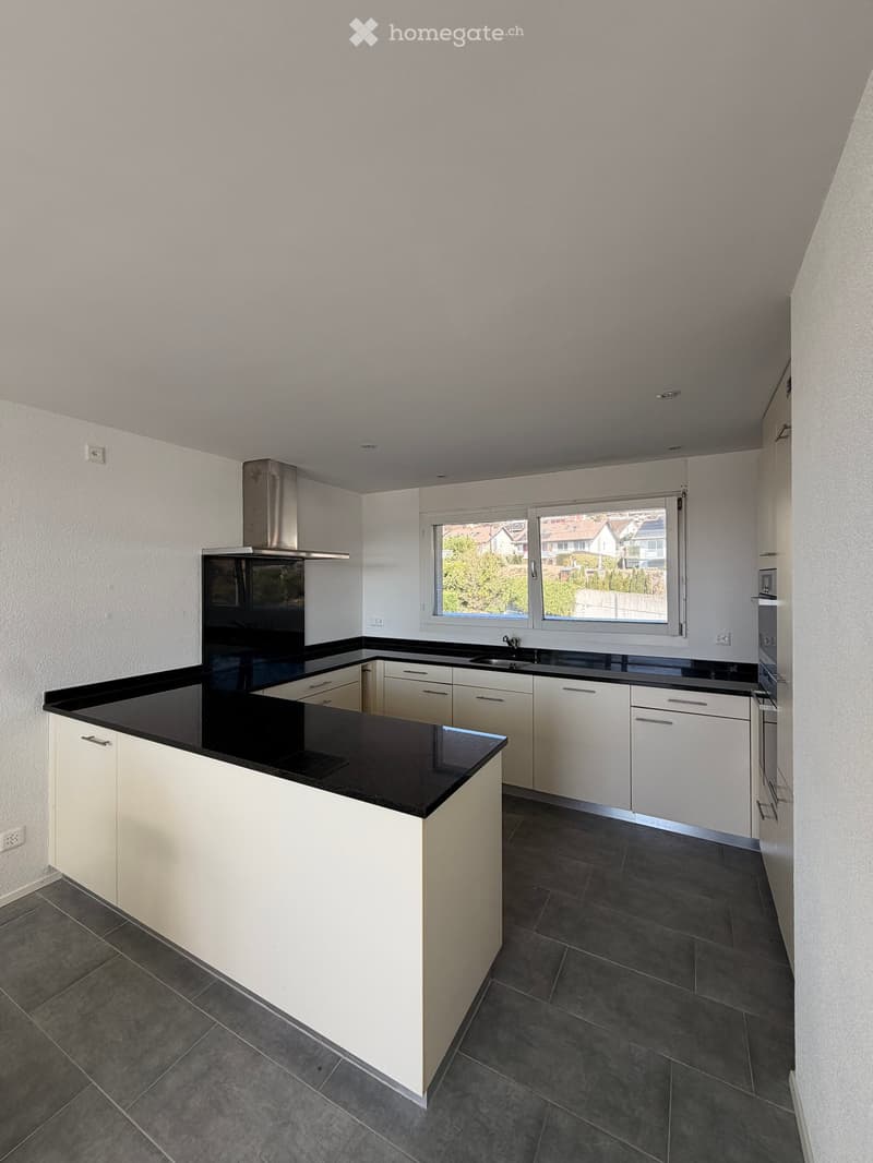 Modern kitchen with white cabinets, black countertops, and a central island. Features a stainless steel range hood and a large window overlooking a residential area.