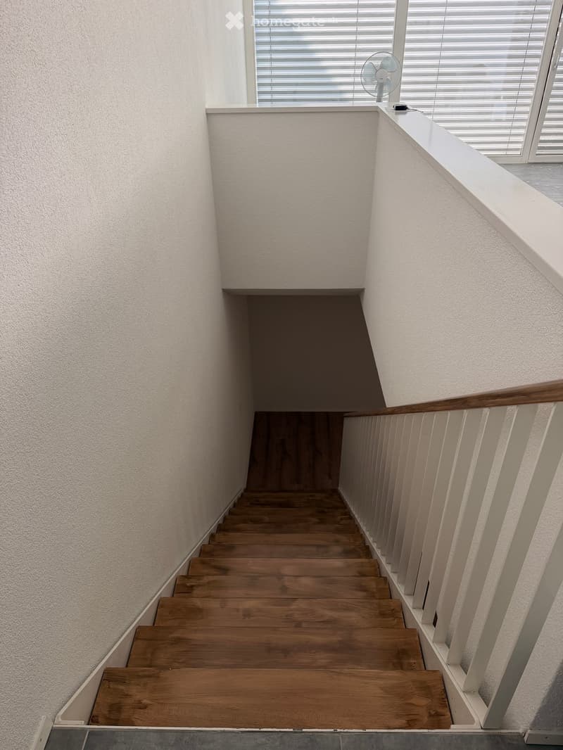Wooden staircase with white walls and a wooden handrail, leading up to a landing area with a window and a fan on the windowsill.