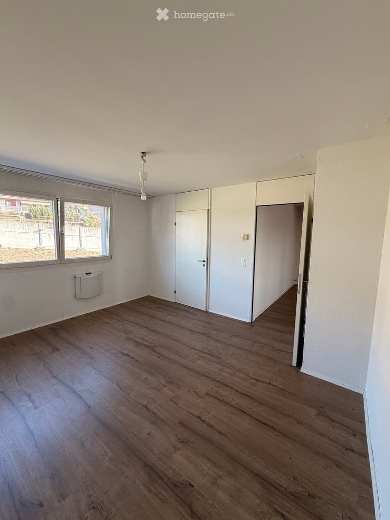 Empty room with wooden flooring, white walls, and large windows. Features a built-in closet and a small trash bin near the window.