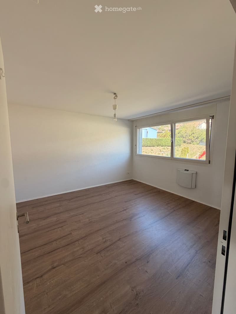 Empty room with wooden flooring, white walls, and a window with a view of greenery outside.
