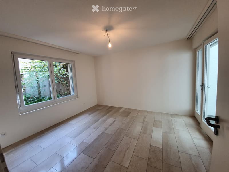 Empty room with light-colored tiled floor, white walls, and large windows showing greenery outside. No furniture or equipment present.