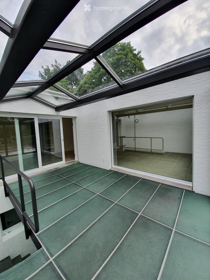 Green tiled balcony with large glass roof sections, white walls, and glass doors leading to interior spaces.