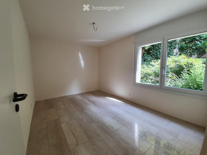 Empty room with tiled floor, white walls, and large window showing greenery outside.