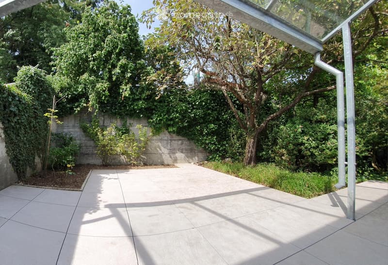 A modern outdoor patio with large tiled flooring, surrounded by lush greenery and trees, featuring a glass-roofed structure on one side.