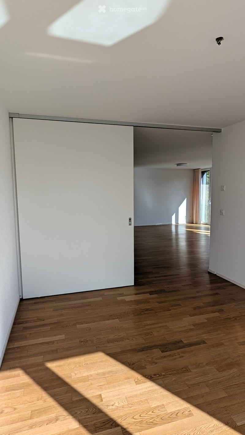 Empty room with wooden flooring and white walls, featuring a sliding door and natural light from a window at the end of the hallway.