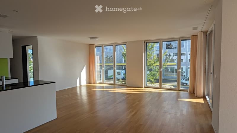 Bright, open room with wooden flooring, large windows, and a small kitchen counter area. No furniture or appliances are present.