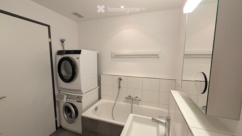 Modern bathroom with a bathtub, sink, mirror, and stacked washing machine and dryer unit.