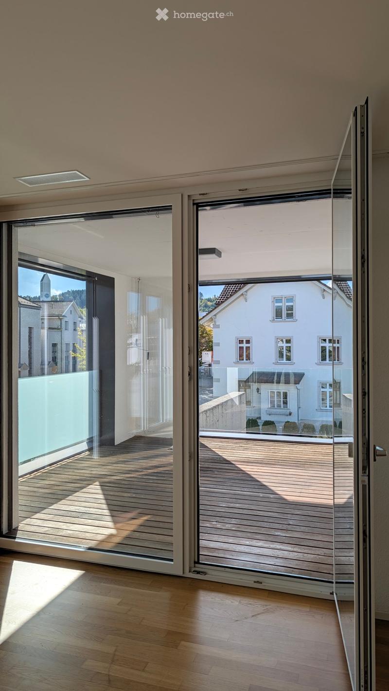 Modern interior with large glass sliding doors leading to a wooden deck, offering a view of a residential area with houses and trees.