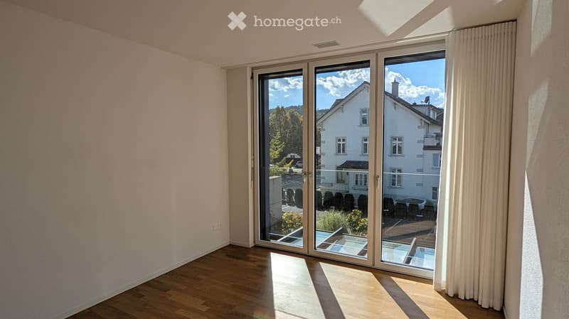 Empty room with wooden flooring, large sliding glass doors leading to a balcony, and a view of a residential area with trees and a house.