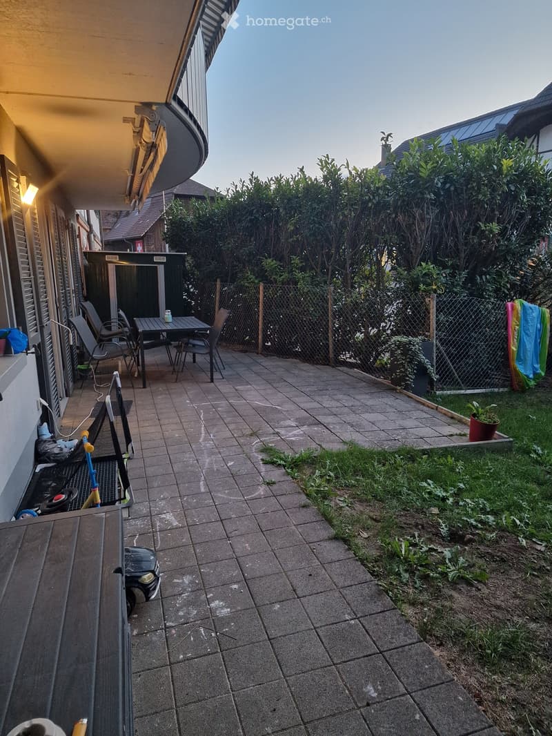 Outdoor patio area with tiled floor, small table and chairs, fenced garden with greenery, and a small toy car.