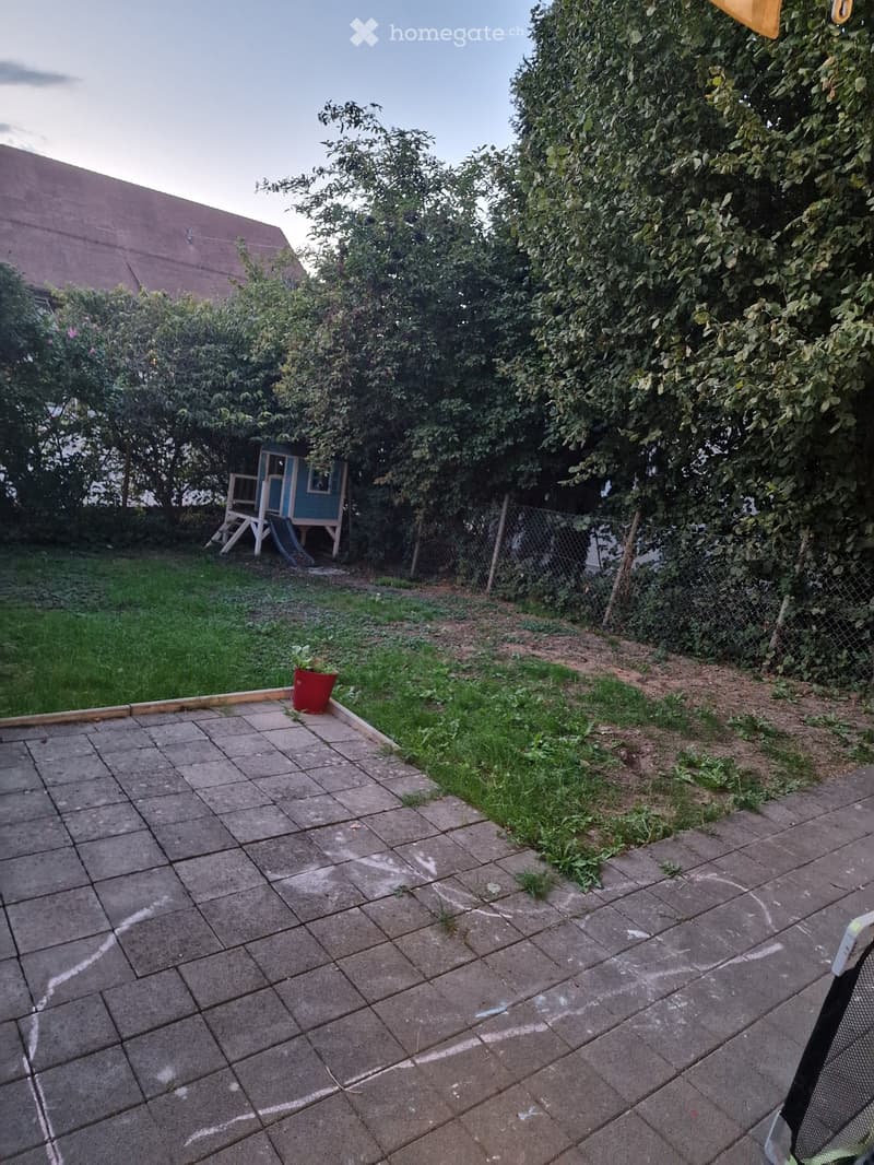 Backyard with a paved patio area, grassy lawn, small playhouse, and a red flower pot. The area is enclosed by a chain-link fence and surrounded by trees and shrubs.