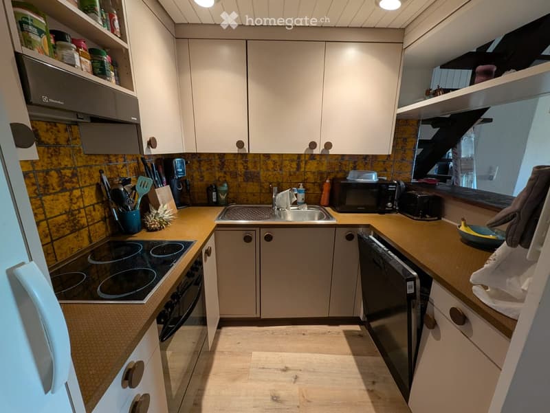 Modern kitchen with light-colored cabinets, wooden countertops, and stainless steel appliances including a refrigerator, oven, dishwasher, microwave, and toaster. The backsplash features a yellow and black tile design. Kitchen utensils and a coffee maker