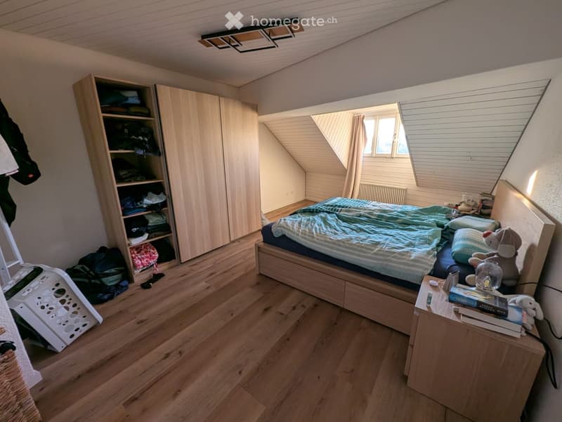 Cozy attic bedroom with wooden floors, slanted ceilings, and large windows. Features a double bed with blue bedding, a nightstand with books and a toy, a wardrobe, and a laundry basket on the floor.