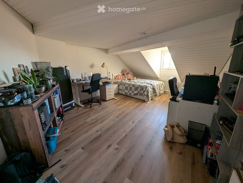 Cozy attic bedroom with wooden flooring, slanted ceilings, and large window. Features a bed with checkered bedding, desk with chair, shelves with books and plants, and a TV on a stand.