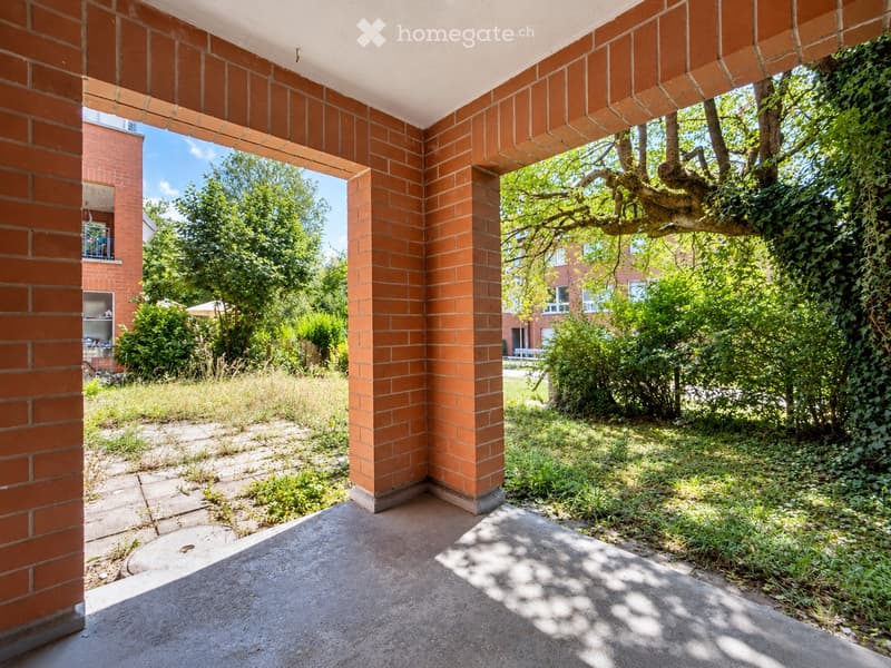 An outdoor patio area with brick columns and a concrete floor, surrounded by greenery and trees, with a view of neighboring buildings and a clear blue sky.