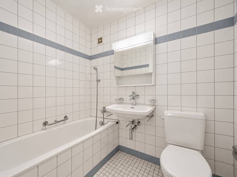 Modern bathroom with white tiled walls and blue accents, featuring a bathtub, toilet, sink, and a mirror with built-in shelf.