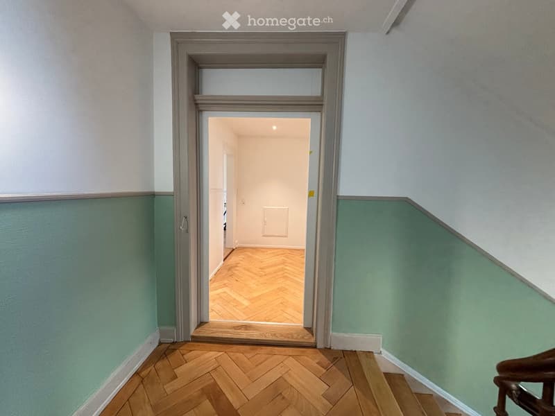 A narrow hallway with wooden herringbone flooring, light green walls, and a door leading to another room with white walls and a visible light fixture.