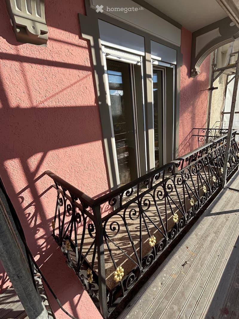 Balcony with ornate metal railing, pink building facade, and glass door leading to interior.