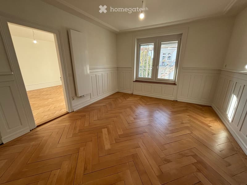 Empty room with herringbone wooden flooring, white walls, and large windows with a view of a building and greenery outside.