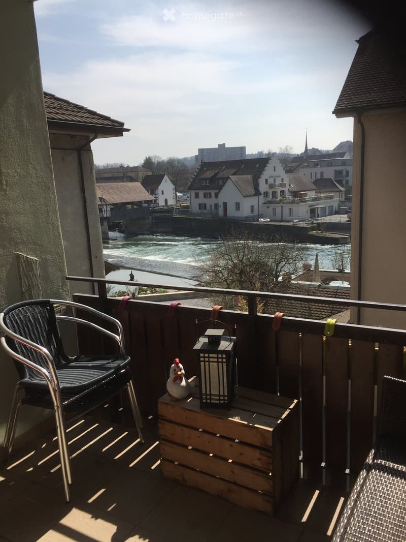 Balcony with a view of a river and town, featuring a wooden crate table, lantern, and decorative chicken figurine.