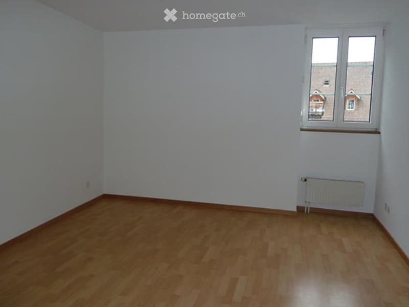 Completely empty room with white walls, wooden floor, and a single window with a radiator below it.