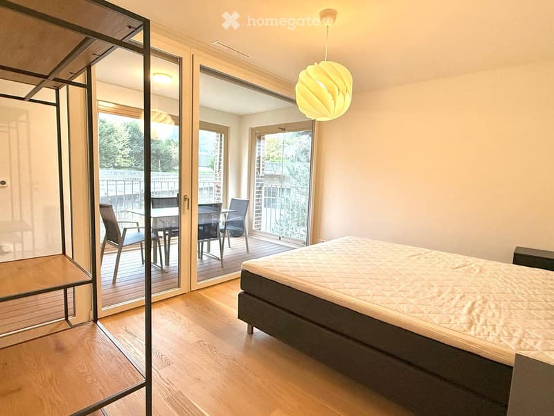 Modern bedroom with wooden flooring, large bed, pendant light, and sliding glass doors leading to a balcony with outdoor furniture.