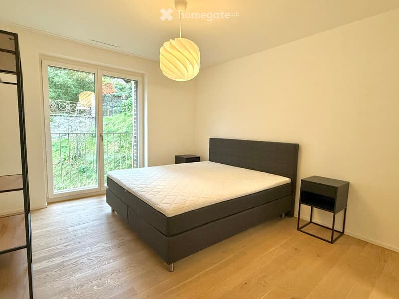 Modern bedroom with a large bed, nightstand, and pendant light. Large sliding glass doors lead to a balcony with greenery visible outside.