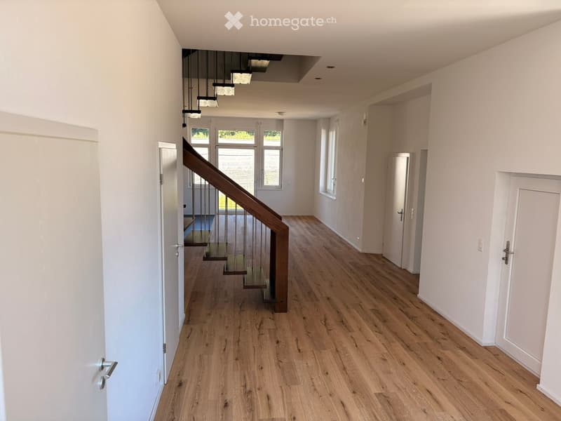 Bright hallway with wooden flooring, white walls, and a staircase leading upwards. Natural light streams in from windows at the far end.