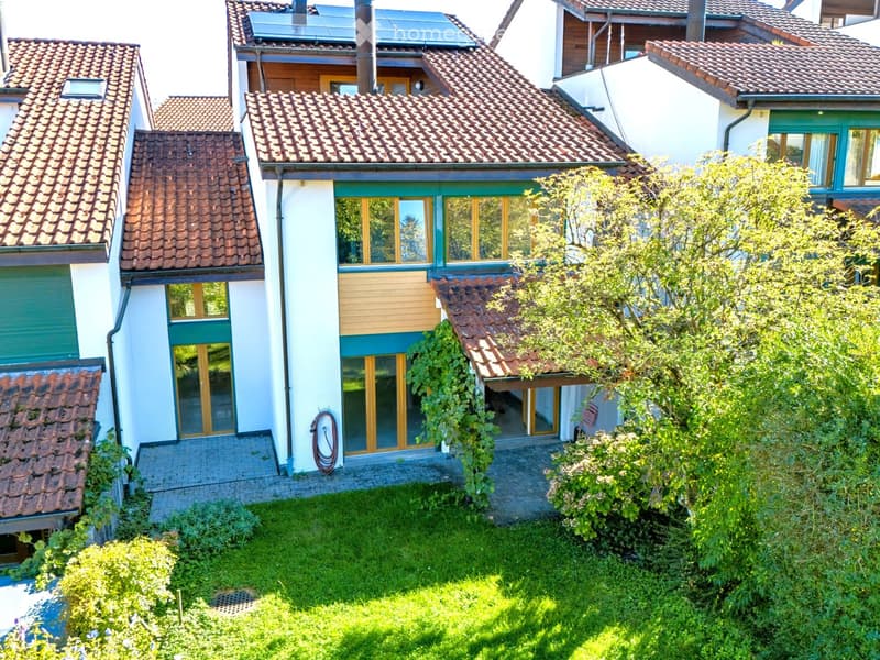 A two-story house with a tiled roof, large windows, and a well-maintained garden. The house features a modern design with light-colored walls and green accents.
