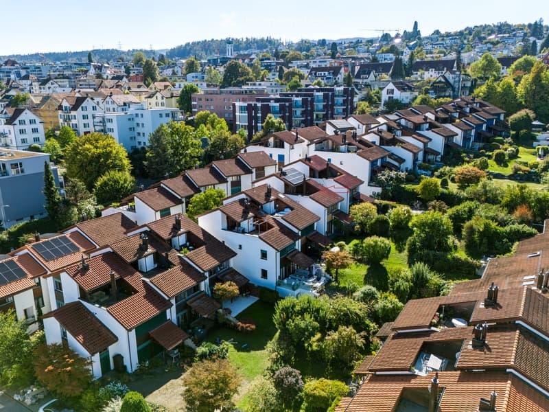 Aerial view of a suburban neighborhood with multiple houses featuring red-tiled roofs, surrounded by greenery and trees. The area includes a mix of residential buildings and landscaped gardens.