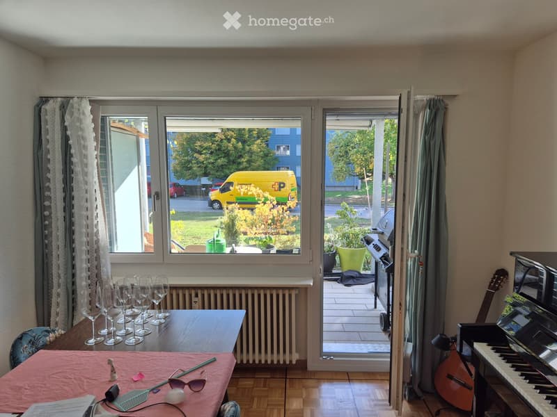 Bright room with wooden flooring, large windows, and a balcony. Features a dining table with wine glasses, a chair, a guitar, a piano, and a curtain.