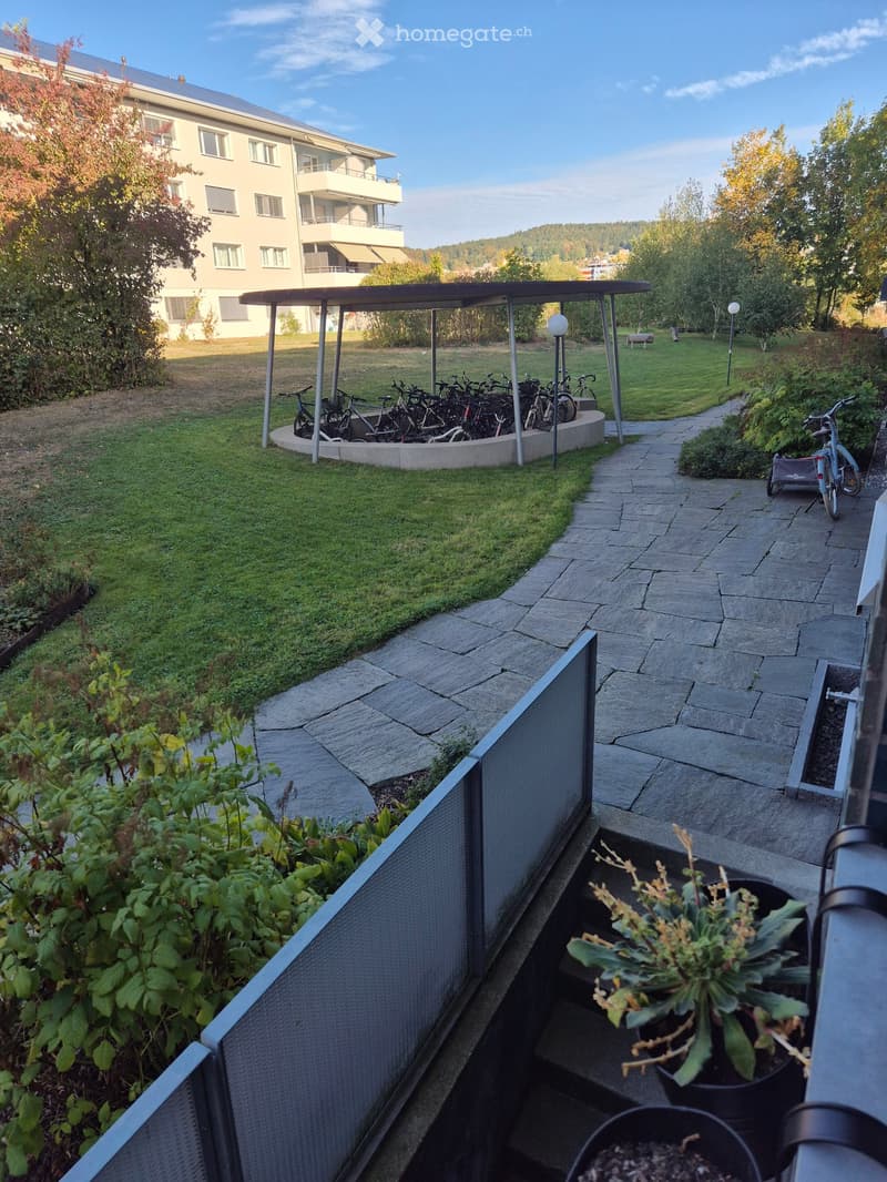 Balcony with potted plants, stone steps, metal railing, and a view of a grassy area with bicycles and a covered shelter.