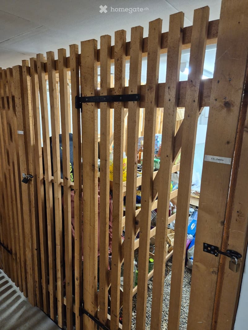 A wooden fence with vertical slats and metal latches separates a hallway from a cluttered storage area. The hallway has a staircase on the left and a sign reading "3 Zi. - Patere" on the right.