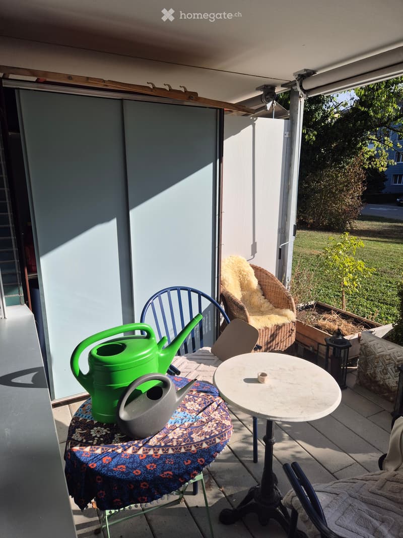 Balcony with a small table, chairs, a wicker chair with a cushion, and a green watering can on a covered surface.