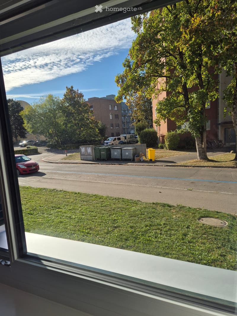 View from a balcony showing a street with parked cars, trash bins, and buildings in the background.