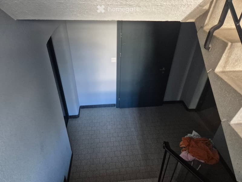 A narrow hallway with tiled floor, light blue walls, and a closed door at the end. A metal railing is visible on the right side, and some clothes are placed near the railing.