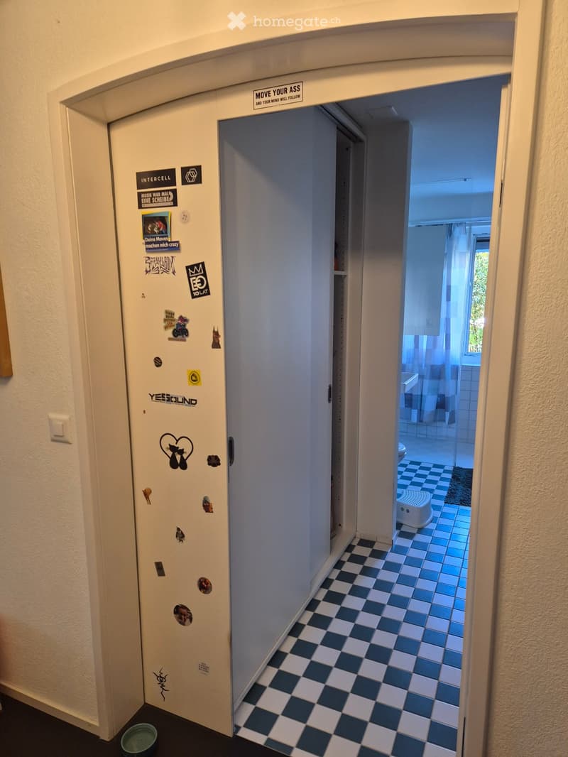 A narrow hallway with checkered floor tiles, leading to a bathroom with blue tiles and a small stool. The hallway walls are adorned with various stickers and signs, including one that reads "MOVE YOUR ASS AND YOUR MIND WILL FOLLOW".