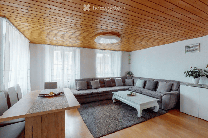 Bright living room with wooden ceiling, large windows, grey sectional sofa, white coffee table, dining table with chairs, and a small plant on a side table.